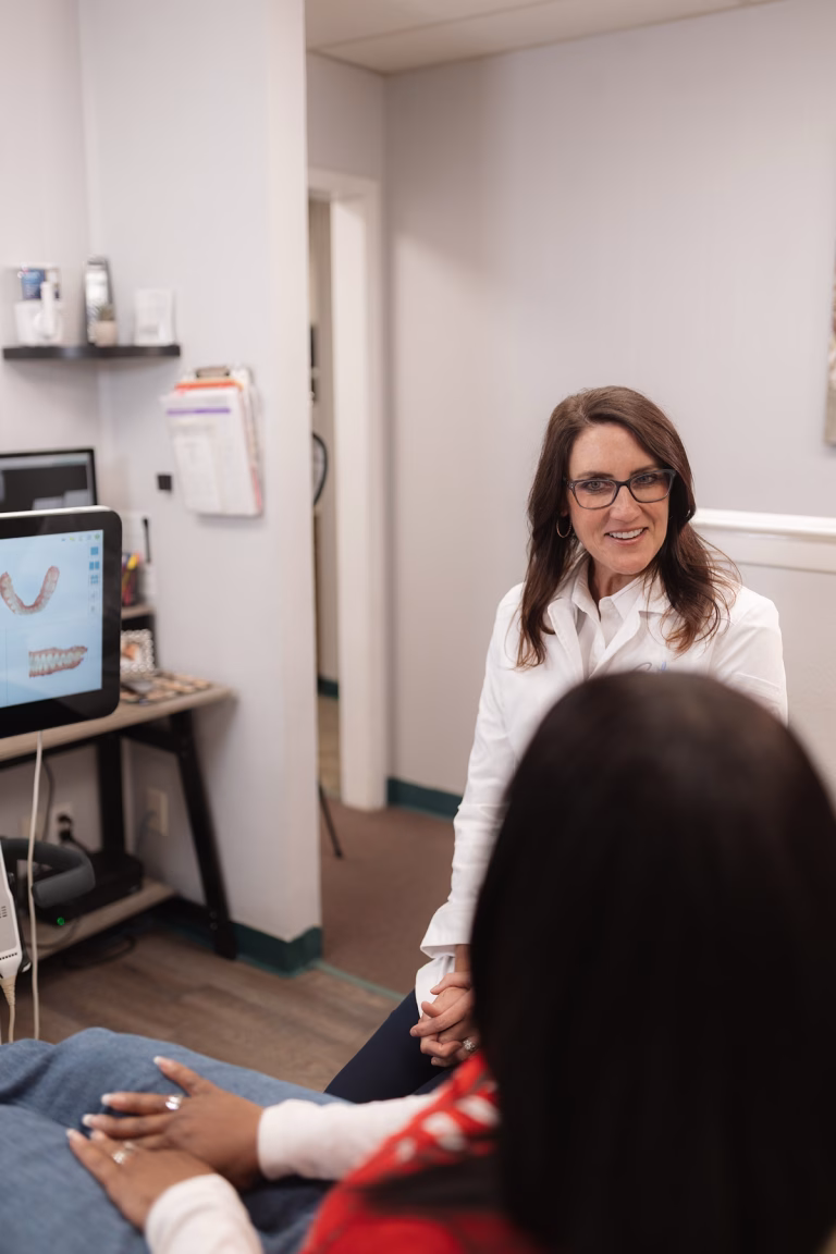 A smiling dentist in a white coat with a patient in an exam room. A dental monitor displays images of teeth, conveying a welcoming and professional atmosphere.