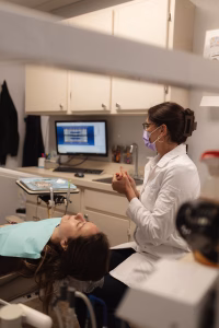 Dentist wearing glasses and a mask talks to a patient in the chair. A monitor shows dental X-rays. The setting is a clean, professional dental office.