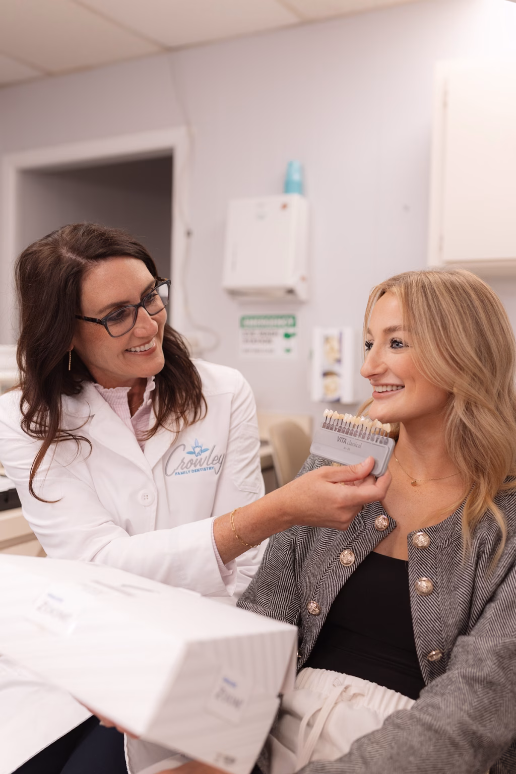 A smiling dentist holds a shade guide, matching teeth color for a patient seated nearby. Both women are smiling, creating a warm and friendly atmosphere.