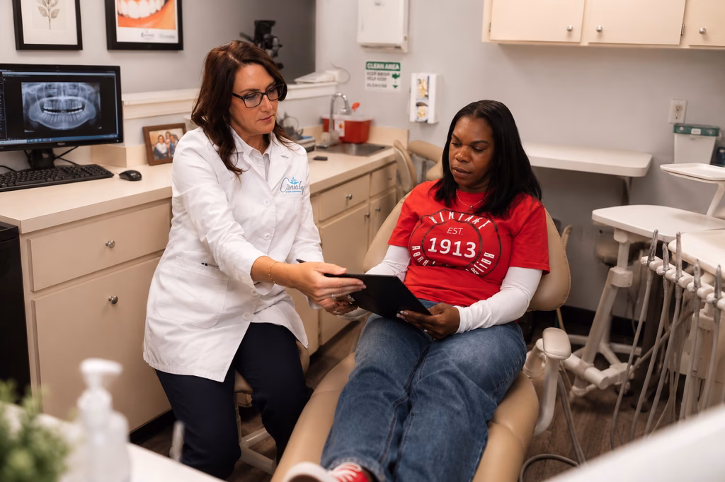 A dental professional in a white coat shows a tablet to a patient in a red shirt sitting in a dentist's chair. The setting is a modern dental office.